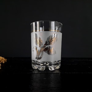 May include: A frosted glass tumbler with a gold leaf design. The glass has a gold band around the top and a gold leaf design on the front.