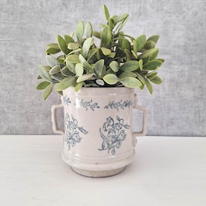 May include: A cream-colored ceramic vessel with two side handles, featuring a blue floral pattern and a crackled glaze finish. The vessel is filled with a cluster of green artificial leaves that extend outward from the top opening.