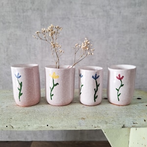 May include: Four pink ceramic cups with hand-painted floral designs. Each cup features a different colored flower: blue, yellow, blue, and red. The cups are arranged on a distressed, light green wooden surface.