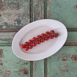 May include: A smooth, oval-shaped white ceramic serving platter holding a single vine of bright red cherry tomatoes. The platter features a clean, minimalist design with a glossy finish, providing a stark contrast to the vibrant red tomatoes.