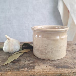 Peut inclure: Un pot en céramique cylindrique avec une glaçure beige brillante et un bord lisse. À côté du récipient se trouvent une gousse d'ail blanche et deux feuilles de laurier séchées, illustrant un contenant de cuisine simple et fonctionnel.