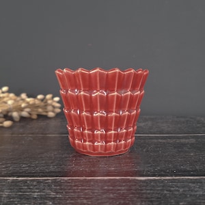 Puede incluir: Una pequeña maceta de cerámica roja con un diseño geométrico texturizado. La maceta tiene un borde ancho y festoneado y un acabado brillante. El recipiente es adecuado para plantas pequeñas o suculentas. El fondo es gris oscuro.