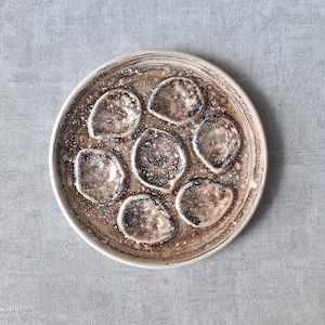 May include: A round, ceramic oyster plate with seven individual wells. The plate features a speckled, brown and cream color scheme with a textured surface. The wells are outlined in dark brown.