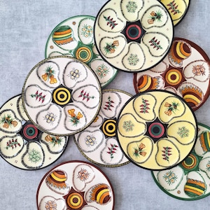 Könnte beinhalten: Eine Sammlung runder Austernplatten aus Keramik, die jeweils sechs muschelförmige Vertiefungen um eine zentrale kreisförmige Vertiefung aufweisen. Die Platten zeigen verschiedene handbemalte Blumen- und geometrische Muster in Gelb, Grün, Braun und Weiß.