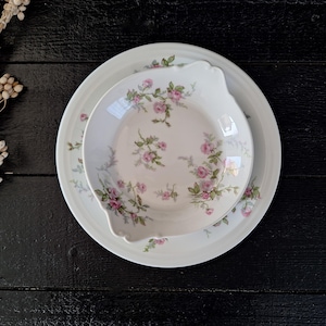 May include: A white porcelain bowl and plate set with a floral pattern of pink roses and green leaves. The set is on a black wooden surface.