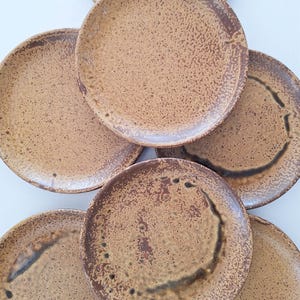 Peut inclure: Ensemble de six assiettes en céramique à surface texturée et mouchetée. Les assiettes présentent une palette de couleurs chaudes et terreuses, avec un centre brun clair et un bord brun plus foncé. Les assiettes sont rondes.