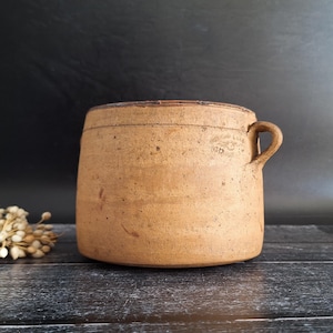 以下が含まれることがあります： 単一のハンドルが付いた、素朴な円筒形の炻器の壺。 アンティークの壺は、暖かく土のような茶色で、わずかにテクスチャ加工された表面です。 ハンドルの近くに小さな、色あせたエンブレムが見えます。 壺は暗い木製の表面に置かれています。