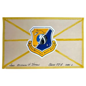 Può includere: Una busta beige con nastro adesivo giallo e un emblema colorato. L'emblema presenta uno scudo con un globo, un satellite e le parole "Defense Meteorological Satellite Program". Il testo scritto a mano include nomi e "Class 77-B".