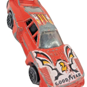 Könnte beinhalten: Ein rotes Spielzeug-Rennauto im Vintage-Stil mit Spoiler und schwarzen Rädern. Das Auto hat weiße und schwarze Grafiken mit der Nummer "2" und dem "GOOD YEAR"-Logo. Der Lack weist Gebrauchsspuren auf.