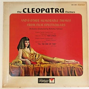 May include: Vintage vinyl record album cover featuring the Cleopatra themes. The cover has a red background with the album title and a picture of a woman in a gold dress. The album includes themes from film spectaculars.