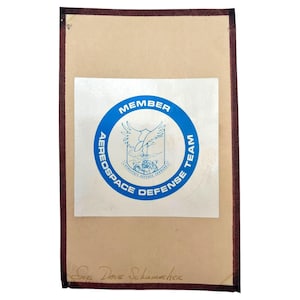 Puede incluir: Un documento rectangular de papel color canela con un emblema circular azul y blanco centrado. El emblema muestra un águila sobre un globo terráqueo con el texto MEMBER AEROOSPACE DEFENSE TEAM. La parte inferior tiene un nombre escrito a mano.