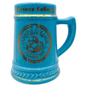 May include: A light blue ceramic beer stein with a handle. The stein has gold trim and a gold emblem with the words "Parsons College" and "Founded 1875". The stein is a classic shape and design.