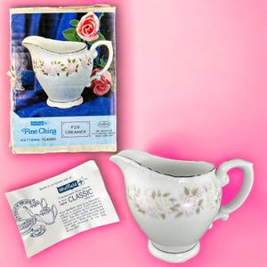 May include: A white porcelain creamer with a floral rose design and a silver trim. The creamer has a handle and a spout. The packaging reads "Sheffield Fine China" and "Classic" pattern. The creamer is approximately 5 inches tall.