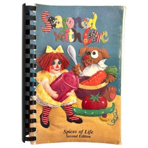 Op de afbeelding: Een spiraalgebonden kookboek getiteld "Seasoned with Love" met een blauwe kaft. De kaft toont een Raggedy Ann pop en een hond die eten bereidt. De tekst "Spices of Life, Second Edition" staat onderaan.