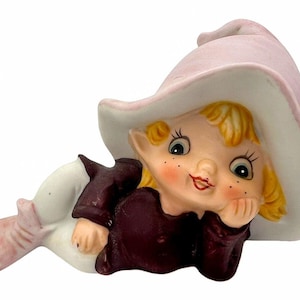 Puede incluir: Una figurita de cerámica de una niña elfo con cabello rubio, que lleva un sombrero rosa y una túnica burdeos. Está tumbada de lado con la cabeza apoyada en la mano.