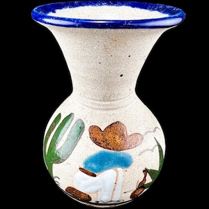 May include: A ceramic vase with a speckled off-white finish and a vibrant blue rim. The body features a hand-painted scene of a figure wearing a brown hat and blue shirt, accompanied by green cactus illustrations against the light-colored base.