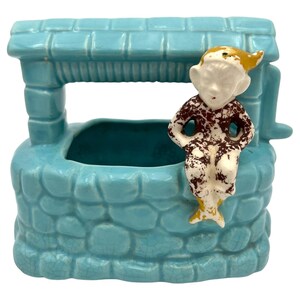 May include: A turquoise ceramic wishing well planter with a seated elf figurine. The well has a textured stone design and a small roof. The elf has a white face, brown and white patterned clothing, and a yellow hat.