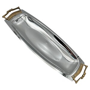 May include: A long, rectangular serving tray with a polished, reflective silver-tone finish. The tray features two ornate, brass-colored metal handles on each end, characterized by a textured, cross-hatched pattern and curved, decorative edges.