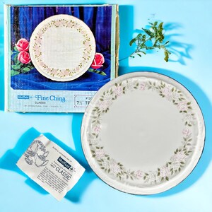 May include: A white fine china plate with a floral rose design and a 7.5-inch diameter. The plate is part of a set, with a matching box featuring roses. The plate has a black rim and a delicate floral border.