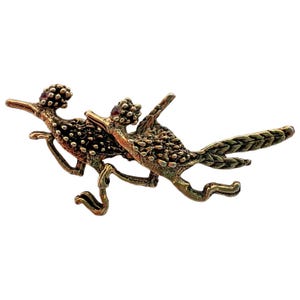 May include: A metallic bronze-toned brooch featuring two roadrunners in a running pose. The birds have textured bodies, long tails, and small red stones set into their crests. The design captures the birds in motion with extended legs.