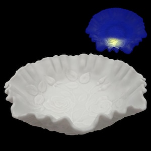 May include: A white, ruffled-edge bowl with embossed rose and leaf designs. The bowl has a scalloped rim and a shallow, rounded shape. The background is black, highlighting the bowl's details.