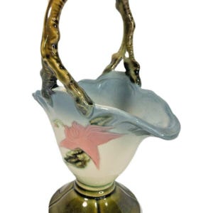 以下が含まれることがあります： 持ち手が付いたセラミックバスケット。水色の縁と、ピンクの花柄で飾られた白いボディが特徴です。ベースは緑色で、持ち手は茶色で枝に似ています。この装飾品はフラワーアレンジメントに適しています。