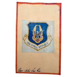 May include: A vintage Air Force Reserve emblem, featuring a blue shield with a gold border and a central eagle design. The emblem is set against a tan background with the words "Air Force Reserve" on a banner. The image is framed with a red border.