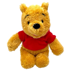 Może przedstawiać: Pluszowa zabawka Kubusia Puchatka, z jasnożółtym korpusem, czarnymi oczami i czarnym nosem. Pluszak ma na sobie jasnoczerwoną koszulkę. Zabawka jest wykonana z miękkiego, puszystego materiału.