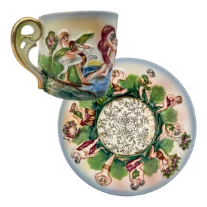 May include: A vintage teacup and saucer set. The cup and saucer are decorated with relief figures and a floral design. The cup has a gold-colored handle and rim. The saucer has a light blue rim and a gold-colored center.