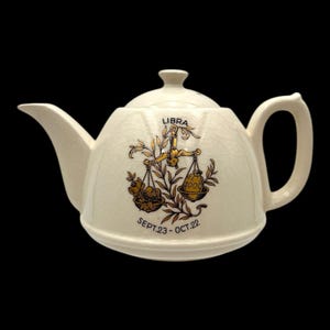 Puede incluir: Tetera de cerámica color crema con tapa, boquilla y asa. La tetera presenta un símbolo del zodiaco Libra dorado con el texto "LIBRA" y "SEPT.23 - OCT.22". Fondo negro.