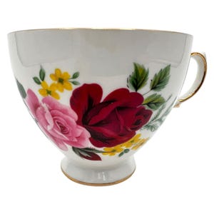 May include: White teacup with a gold rim and handle, decorated with pink and red roses, yellow flowers, and green leaves. The floral design is on the front of the cup. The teacup is a vintage style.