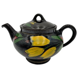 Op de afbeelding: Zwarte keramische theepot met deksel, handvat en tuit. De theepot is versierd met gele tulpachtige bloemen, groene bladeren en rode accenten. Het handvat en de tuit zijn zwart.