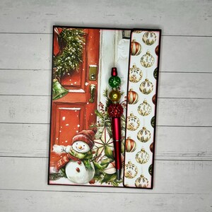May include: A rectangular notepad featuring a festive illustration of a snowman standing before a red door with a wreath, paired with a red ballpoint pen adorned with stacked green, gold, and red textured beads on the top end of the pen barrel.