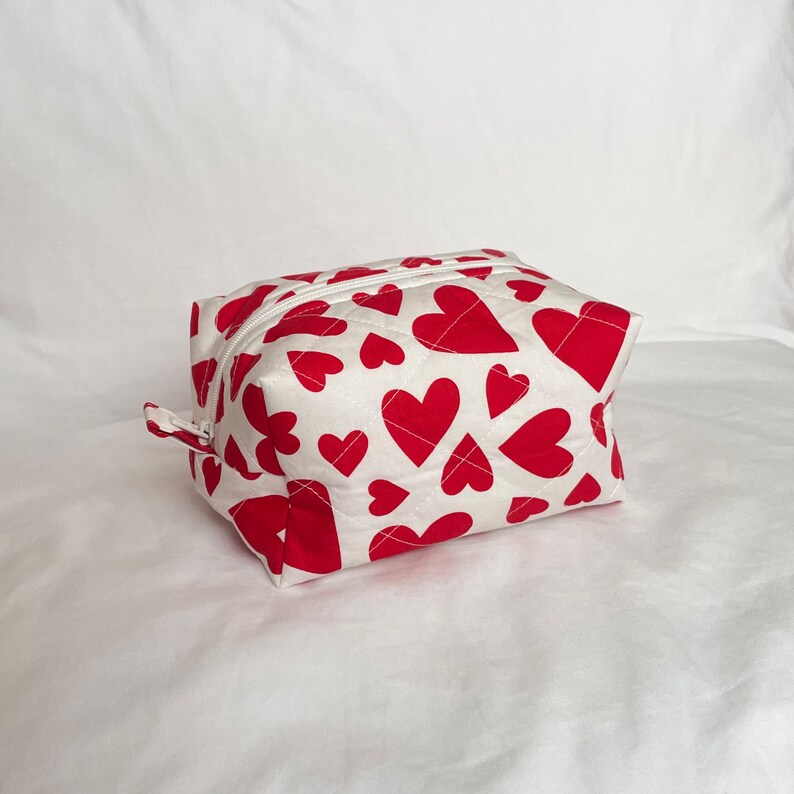 May include: A white quilted cosmetic bag with a pattern of red hearts. The bag has a zipper closure and a small fabric loop on the side. The hearts vary in size and are evenly spaced across the surface.