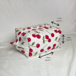 May include: A white quilted cosmetic bag with a cherry pattern. The bag is rectangular, measuring 6.25 inches long, 4.25 inches wide, and 3.5 inches high. It has a white zipper and a cherry fabric pull.