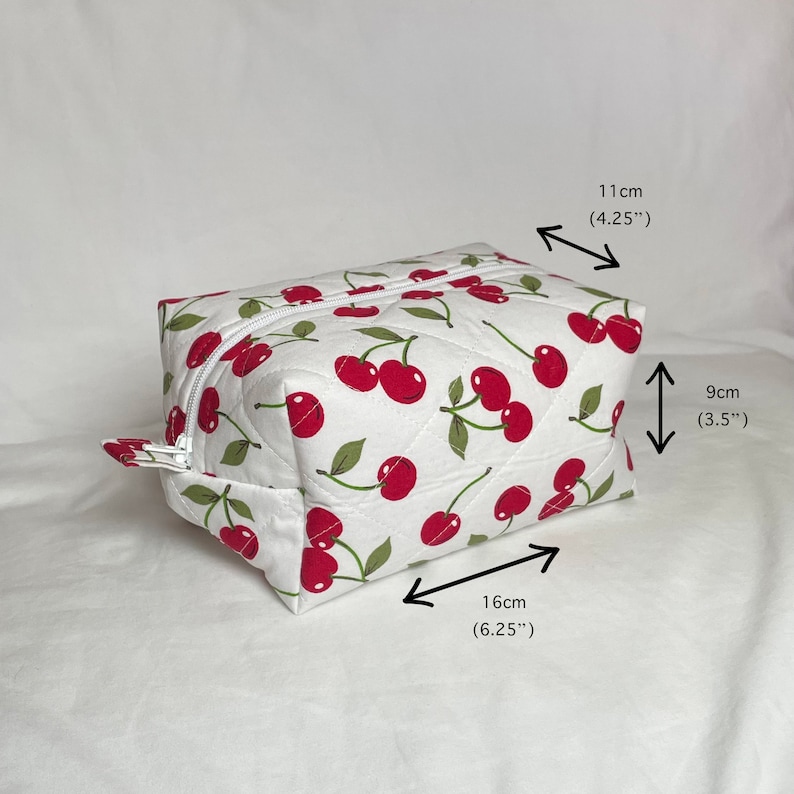 May include: A white quilted cosmetic bag with a cherry pattern. The bag is rectangular, measuring 6.25 inches long, 4.25 inches wide, and 3.5 inches high. It has a white zipper and a cherry fabric pull.