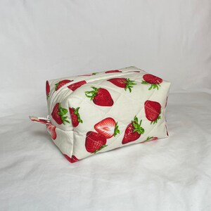 May include: A quilted, rectangular cosmetic bag with a white background and a repeating pattern of red strawberries with green leaves. The bag has a zipper closure and a small fabric pull.