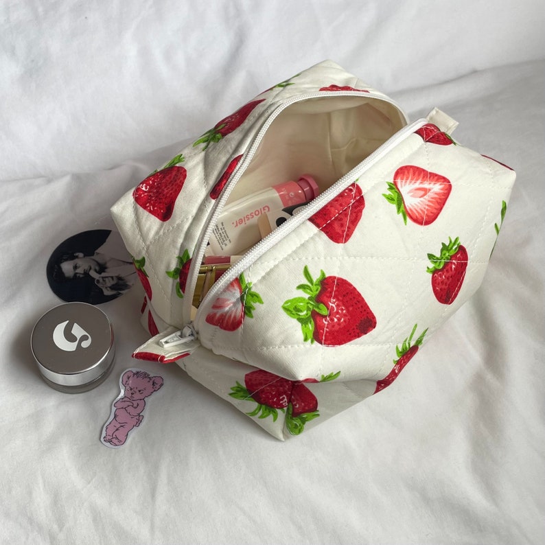May include: A quilted cosmetic bag with a white background and a repeating pattern of red strawberries with green leaves. The bag is open, revealing a pink lip gloss and other beauty products. A silver container and stickers are next to the bag.