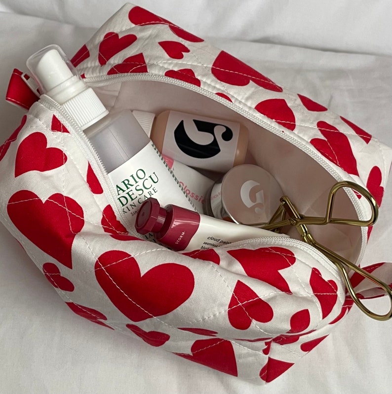 May include: A white cosmetic bag with a red heart pattern, open to reveal various beauty products. The bag contains a spray bottle, tubes, and a gold-colored eyelash curler. The bag is zippered and has a quilted design.