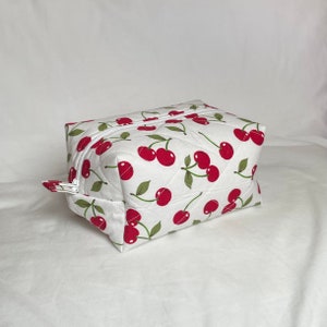 May include: A white quilted cosmetic bag with a repeating pattern of red cherries and green leaves. The bag has a rectangular shape with a zipper closure and a small fabric loop on the side.