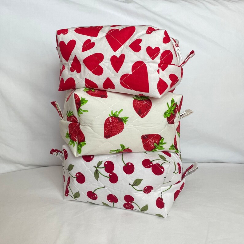 May include: Three quilted cosmetic bags stacked on top of each other. The top bag has a white background with red hearts. The middle bag has a white background with red strawberries. The bottom bag has a white background with red cherries.