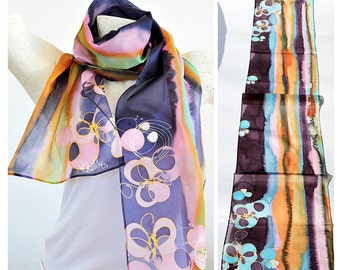 May include: A silk scarf with a colorful abstract design featuring circles and swirls in shades of pink, blue, orange, and yellow. The scarf is hand-painted and has a black background. The scarf is shown on a mannequin and in a close-up view.