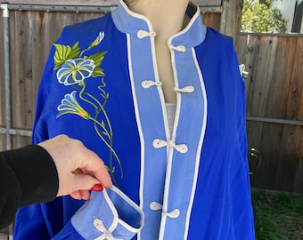 Puede incluir: Una camisa de seda azul con cuello y puños blancos. La camisa tiene un cuello mandarín y está decorada con flores bordadas en la parte delantera. La camisa tiene un ribete blanco y detalles azules.