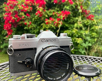 Może przedstawiać: Srebrny aparat fotograficzny Canon FTb 35 mm z obiektywem Canon. Aparat ma czarny skórzany uchwyt i czarny obiektyw. Aparat stoi na czarnym metalowym stole.