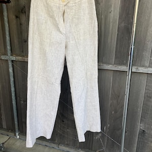 May include: Cream-colored, vertically striped pants hanging on a wooden hanger. The pants have a button closure and a straight leg design. The stripes are a dark gray color.