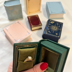 May include: A collection of small, book-shaped ring boxes in various colors including light blue, cream, pink, dark red, and navy blue with gold celestial detailing. One open green box features a velvet cushion holding a gold filigree ring.