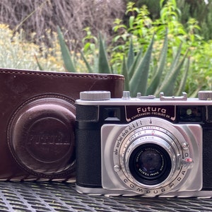 May include: A silver and black 35mm film camera with a leather case. The camera has a Compur-Rapid shutter and a  Zeiss Tessar lens. The camera is labeled "Futura" and "Kamerawerk Freiburg Br."