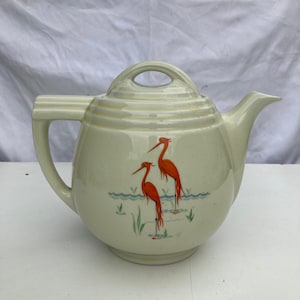 May include: A cream-colored ceramic teapot with a lid and a handle. The teapot has a design of two orange birds standing in a pond with green reeds.