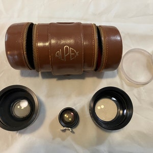 May include: A set of four camera lens filters in a brown leather case. The case has the word "ALPE" embossed on it. The filters are black and have a silver ring around the edge.