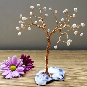 May include: A handcrafted copper wire tree sculpture with white pearl accents, set on a blue and white stone base. The tree has a twisted trunk and delicate branches. Two purple and pink flowers are in the foreground.